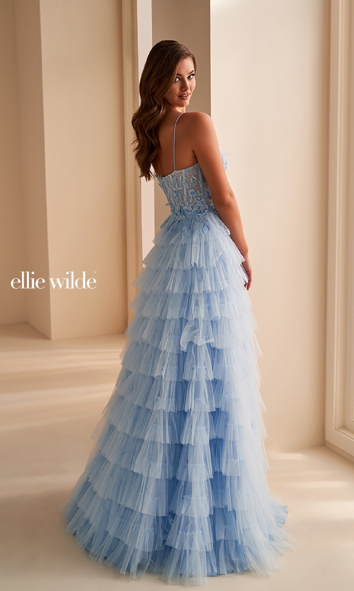 A woman indoors wears the Ellie Wilde EW36223 long prom dress—a light blue, tiered ruffle gown with thin straps and an intricate corset bodice. She looks over her shoulder as